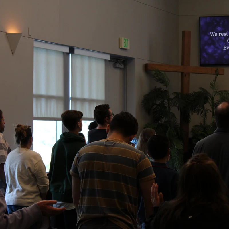A diverse group of people seen from behind, standing and worshiping in a church. A screen displays song lyrics next to a large wooden cross.